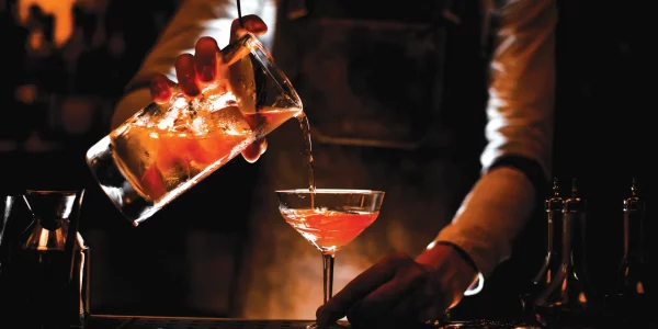 Male bartender in leather apron pouring the alcohol cocktail with steel strainer