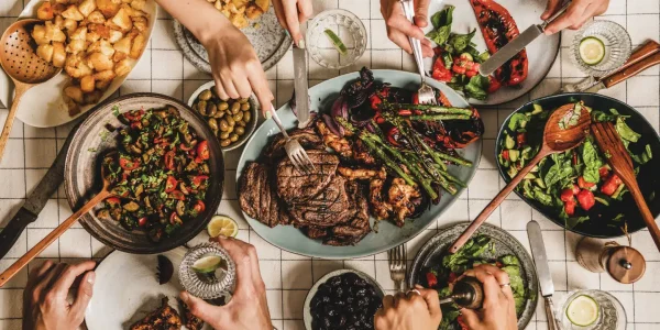 Summer barbeque party. Flat-lay of table with grilled meat, vegetables, salad, roasted potato and peoples hands over checkered white tablecloth, top view. Family gathering, comfort food concept